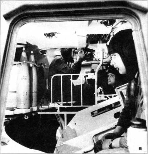 Вид на интерьер САУ «Акация» через открытый бортовой люк. На переднем плане заряжающий (сидит спиной к движению), за ним и за ограждением орудия — наводчик и командир машины.