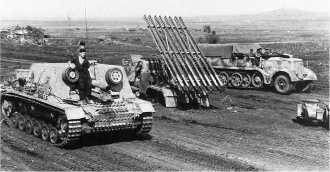Тяжелое самоходное пехотное орудие StulG 33 из состава 23-й танковой дивизии Вермахта выдвигается к передовой. На заднем плане: 18-й тонный полугусеничный тягач Sd.Kfz.9 и разбитая «Катюша» — установка БМ-13Н на шасси Studebaker US6. «Миус-фронт», июль 1943 года.